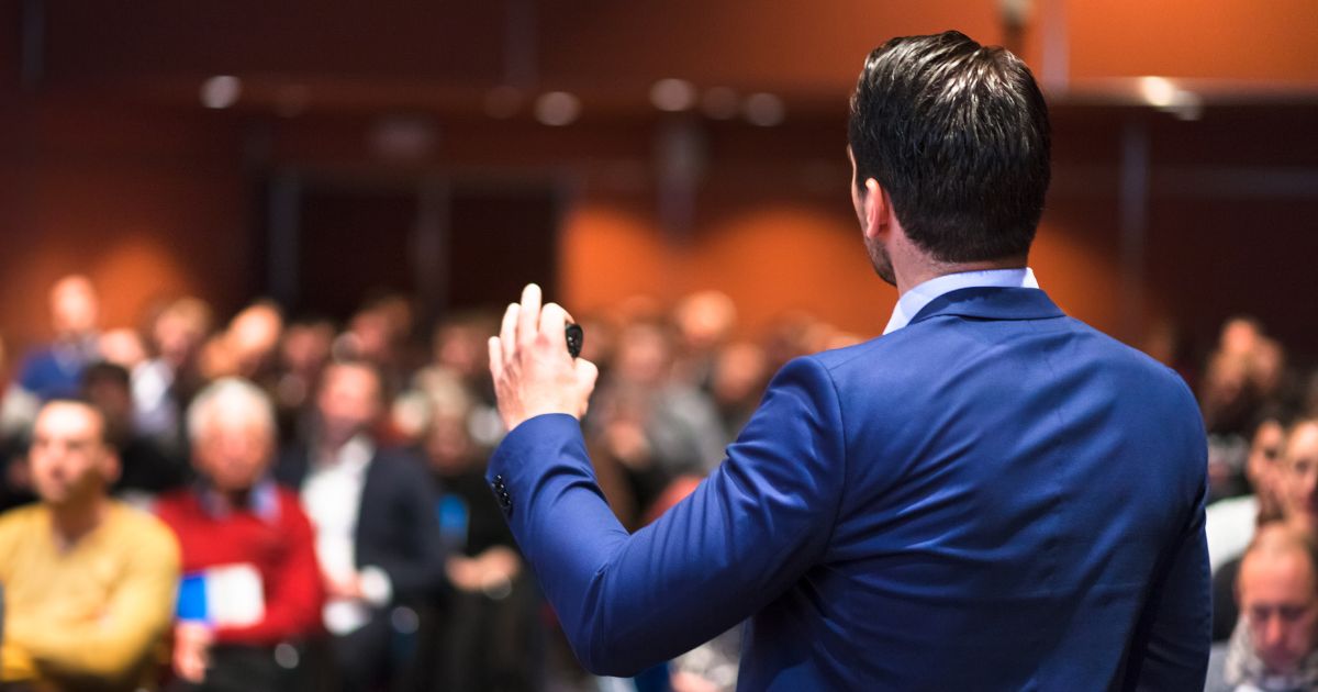 image of a speaker from behind reaching the mic in front of a crowded audience to show how the 10 Best Sales Kickoff Speakers for 2027 Top Keynotes to Drive Revenue